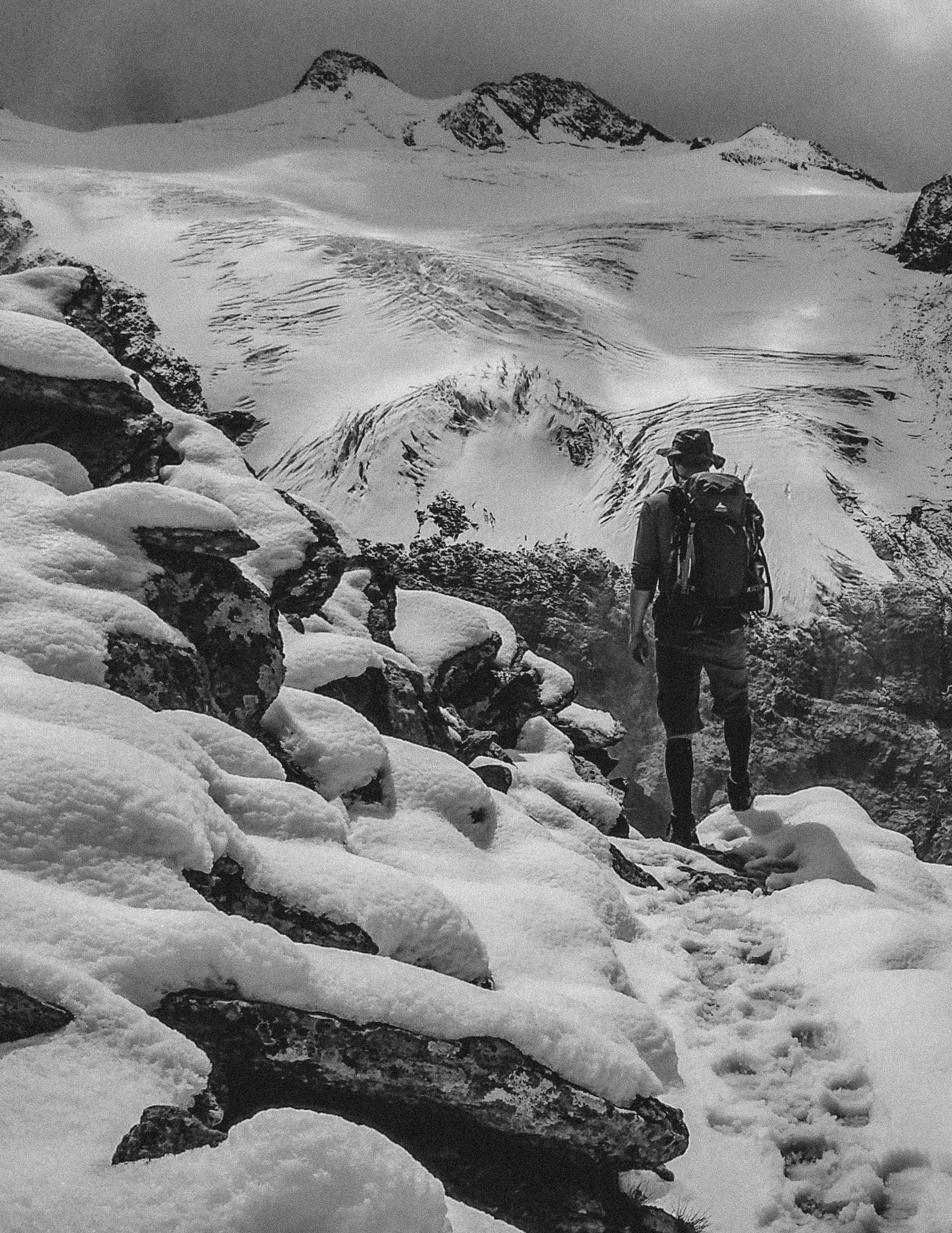 Wanderer mit Rucksack steht auf einem schneebedeckten Weg auf einem Berg und blickt auf einen Gletscher auf der anderen Talseite